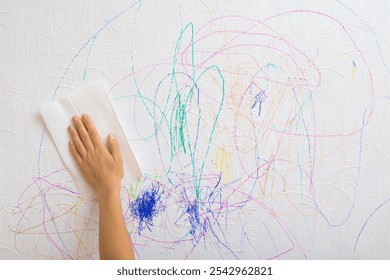 Woman hand holding white dry paper napkin and wiping scratches lines of colorful markers from white wallpaper surface. Mother cleaning wall from draw of naughty little kid. Closeup. Front view. - Powered by Shutterstock