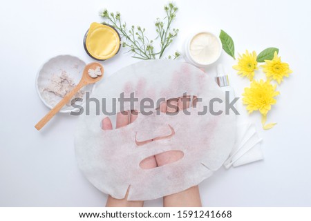 woman hand holding white sheet mask with clay mask wooden spoon yellow flowers and serum skincare cream products on white background. Aerial view