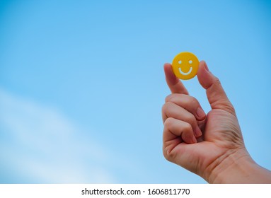 Woman Hand Holding Yellow Emoticon Smiley Face Icon Symbol On Blue Sky Background With Copy Space, For A Positive Mindset In Business Marketing And Beautiful World Peace Concept