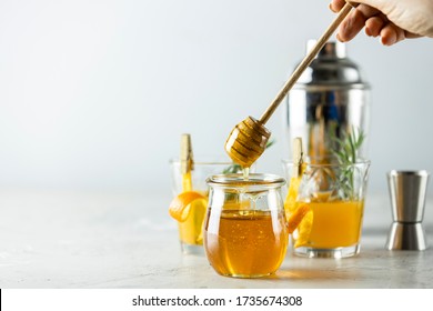 Woman Hand Holds A Spoon For Honey Over Jar In Front Of Two Glasses Of Honey Bourbon Cocktail With Rosemary Syrup Or Homemade Whiskey Sour With Orange Peel And Rosemary Decoration And Bartender Tools