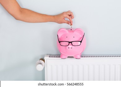 Woman Hand Inserting Coin In Piggy Bank On Radiator To Save On Energy Bill