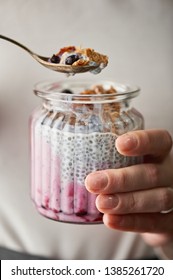 Woman Hand Keeps A Jar Of Glass Full Of Chia Pudding And Metal Spoon Full Of Pudding, Oatmeal Flakes And Black Currant Above The Jar