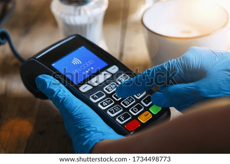 Woman hand with medical latex protective gloves holds payment terminal and enters PIN code at a cafe. Protection for Coronavirus or COVID-19. 
