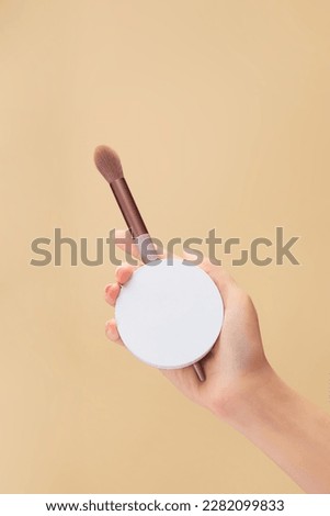 A woman hand model carrying a compact powder case with a blending brush against a beige background. Empty label for product mockup