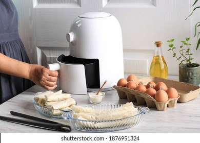 Woman Hand Open Airfryer Tray. A White Deep Fryer or Oil Free Fryer Appliance, Tongs, Clear Baking Dish and Egg Tray are on the Wooden Table in the Kitchen with a Small Plant in the Pot (air fryer)