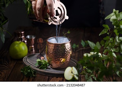 Woman hand pouring Green Apple Irish Mule cocktail cocktail from shaker in copper mug surrounded by ingredients and bar tools on dark wooden table surface
