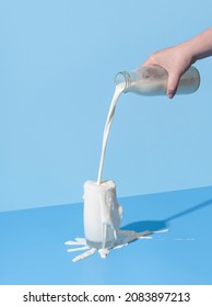 Woman hand pouring milk from a bottle into the glass, minimalist on a blue background. Over-spilling milk on the blue-colored table. - Shutterstock ID 2083897213