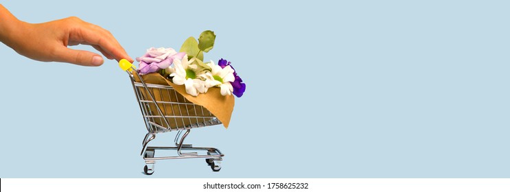 A Woman Hand Pushing And Rolling A Mini Shopping Cart With Flowers On A Blue Background. Spring Shopping Online Concept. Seasonal Sale. Flowers Fast Devilery. Banner No People. Space For Text.