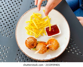 Woman hand putting fried potato into the ketchup on table with small burgers on the plate. Hamburger. Burger. Meat. Small. Food. Snack. Fast Food.  Meal