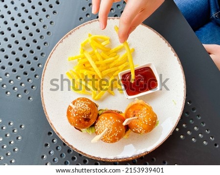 Woman hand putting fried potato into the ketchup on table with small burgers on the plate. Hamburger. Burger. Meat. Small. Food. Snack. Fast Food.  Meal