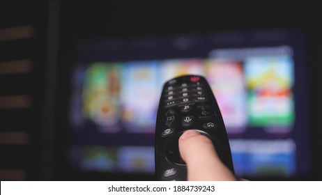 Woman Hand Selects Internet Tv Channels With Remote Control, Close-up. Person Controls TV Using A Modern Remote Control. Girl Watches Smart TV And Uses Black Remote Control. Blurry Tv Scrolls Pages.