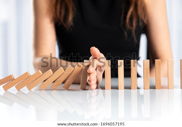 Woman hand stopping falling wooden dominoes effect\
on white solid ground
