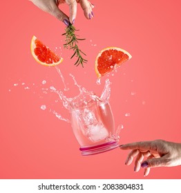 Woman hand support fly glass of grapefruit drink with splash, juice grapefruit slices falling in glass. Cocktail of grapefruit, thyme and rosemary flavor. Summer art food concept on pink background