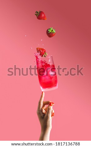 Woman hand support flying glass of strawberry cocktail drink with splash, juice strawberries falling in glass. Summer art food concept on pink background.