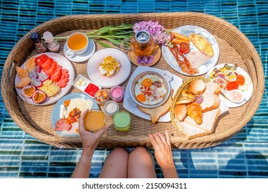 Woman Hand Take Fruit Juice Glass Of Floating Breakfast Tray In Swimming Pool On Hotel Villa Resort. Set With Eggs Benedict, Sunny Eggs, Plate With Tropical Fruits, Bread Basket, Tea Cup And Teapot.