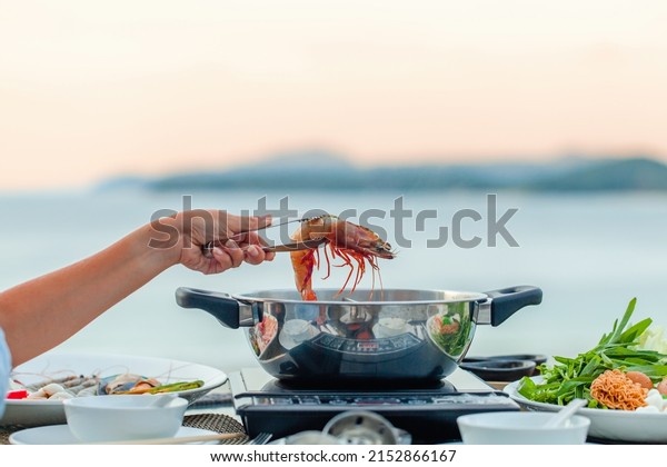 Woman hand take river prawn and put it in\
boiling pot Shabu shabu hotpot style in Japanese restaurant. Shabu\
Shabu dinner near swimming pool. Sukiyaki dish of pork, shrimp,\
seafood, fresh\
vegetables.