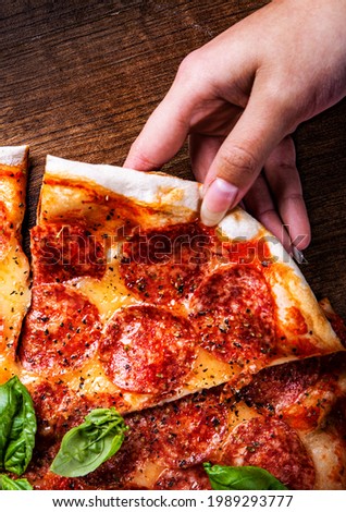 woman Hand takes a slice of Pepperoni Pizza with Mozzarella cheese, salami, Tomatoes, pepper, Spices and Fresh Basil. Italian pizza on wooden table background
