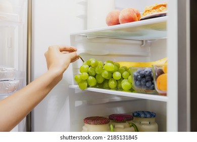 Woman hand taking, grabbing or picks up green bunch of grapes out of open refrigerator shelf or fridge drawer full of fruits, blueberries, bottles with yogurt. Healthy food diet, lifestyle concept - Shutterstock ID 2185131845