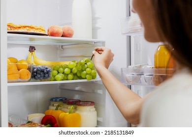 Woman hand taking, grabbing or picks up green bunch of grapes out of open refrigerator shelf or fridge drawer full of fruits, vegetables, banana, peaches, yogurt. Healthy food diet, lifestyle - Shutterstock ID 2185131849