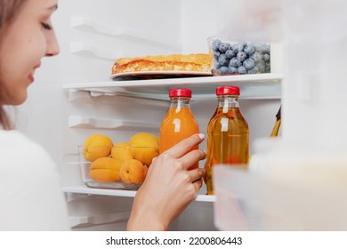 Woman Hand Taking, Grabbing Or Picks Up Juice Glass Bottle Out Of Open Refrigerator Shelf Or Fridge Drawer Full Of Fruits, Vegetables, Banana, Peaches, Yogurt. Healthy Food Diet, Lifestyle Concept