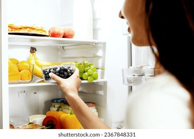 Woman hand taking, grabbing or picks up box of blueberry out of open refrigerator shelf or fridge drawer full of fruits, vegetables, banana, peaches, yogurt. Healthy food diet, lifestyle concept - Shutterstock ID 2367848331