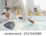 Woman hand touching clear blue water of hot tub on terrace. Female in linen robe sitting on corner of bath enjoying scenic sunset during summer time. Hotel resort, recreation and vacation concepts