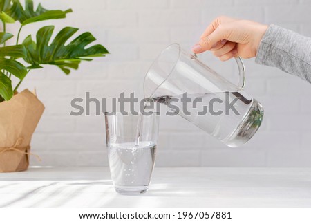 Woman hand with transparent jug fills a glass pure drinking water, creating air bubbles, green plant leaf on white table at home. Freshness, cold refreshing drink, water balance, health, thirst, diet.