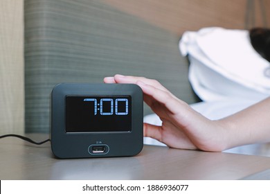 Woman hand turning off black digital alarm clock on the table side bed after awakening in morning at 7.00 o’clock.