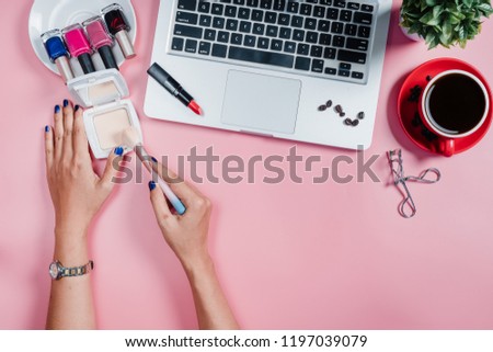 Woman hand use a make-up brush on pink desk with laptop,cup of coffee and cosmetic set.Top view,Flat lay.