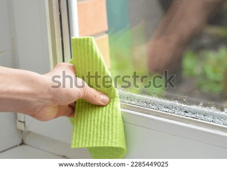 Woman hand wipes off water condensation in room with high humidity level. Home moisture with water condensation on the window glass.