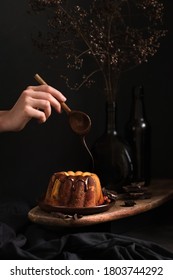 Woman Hand With Wooden Spoon Drizzling Chocolate Icing On Easter Or Christmas Bund Cake, Dark Mood Photography, Dried Flowers On A Background