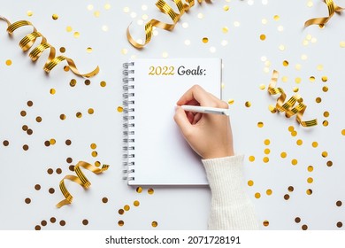 Woman Hand Writing 2022 New Year Resolution Notebook With Gold Festive Decoration On A White Background, Flat Lay Style