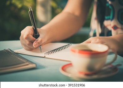 Woman Hand Writing Journal On Small Notebook At Outdoor Area In Cafe With Morning Scene And Vintage Filer Effect