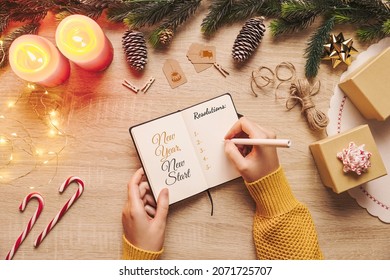 Woman Hand Writing In A New Year Resolution Notebook With Checklist Of Goals And Christmas Rustic Decor Stuff On A Wooden Background. Top View Blank Paper, New Start Concept