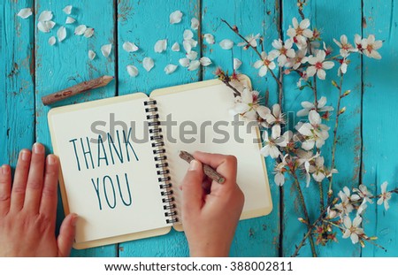 woman hand writing a note with the text thank you on a notebook, over wooden table and cherry blossom flowers