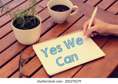 Woman Hand Writing In Notebook Yes We Can On Wooden Table With Coffee, Plant And Glasses. Retro Colors.