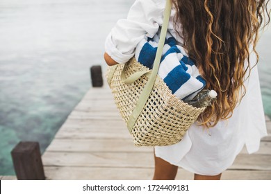 Woman with handmade wicker bag, two beach towels and glass bottle for water going to the beach. View from behind close up. Eco friendly and zero waste concept.