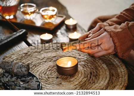 Woman hands burning Palo Santo, before ritual on the table with candles and green plants. Smoke of smudging treats pain and stress, clears negative energy and meditation wooden stick
