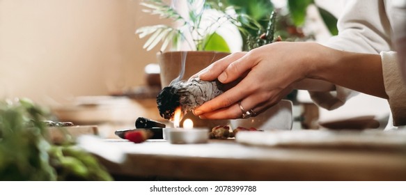 Woman Hands Burning White Sage, Palo Santo Before Ritual On The Table With Candles And Green Plants. Smoke Of Smudging Treats Pain And Stress, Clear Negative Energy