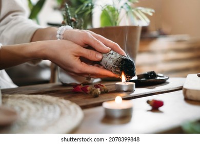 Woman Hands Burning White Sage, Palo Santo Before Ritual On The Table With Candles And Green Plants. Smoke Of Smudging Treats Pain And Stress, Clear Negative Energy