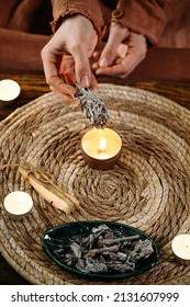 Woman Hands Burning White Sage, Before Ritual On The Table With Candles And Green Plants. Smoke Of Smudging Treats Pain And Stress, Clear Negative Energy And Meditation