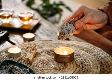 Woman Hands Burning White Sage, Before Ritual On The Table With Candles And Green Plants. Smoke Of Smudging Treats Pain And Stress, Clear Negative Energy And Meditation