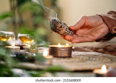 Woman hands burning white sage, before ritual on the table with candles and green plants. Smoke of smudging treats pain and stress, clear negative energy and meditation