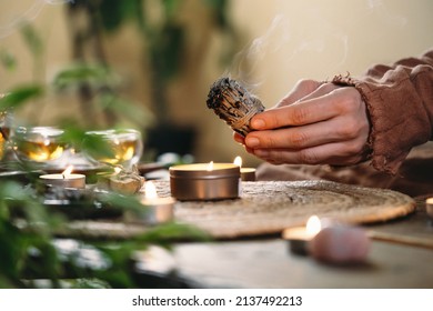 Woman Hands Burning White Sage, Before Ritual On The Table With Candles And Green Plants. Smoke Of Smudging Treats Pain And Stress, Clear Negative Energy And Meditation