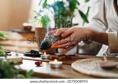 Woman hands burning white sage, palo santo before ritual on the table with candles and green plants. Smoke of smudging treats pain and stress, clear negative energy