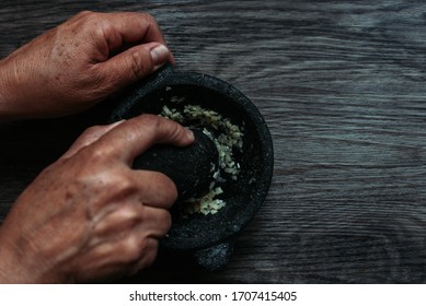 Woman Hands Crushing Garlic With Stone In A Mortar On A Wooden Board, Top View Of Traditional Preparation In A Mortar