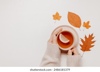 Woman hands hold cup of tea with lemon with autumn leaves at the background on white table, copy space. Woman in sweater with tea. autumn cozy mood. - Powered by Shutterstock