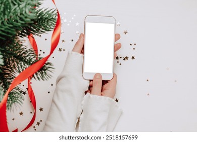 Woman hands hold smartphone with white empty screen with golden confetti , christmas fir tree on wooden table, top view, background for holidays festive website, online shopping and design - Powered by Shutterstock