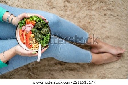 Woman hands holding bowl of plant based food while sitting on the carpet of her living room. Close up caption