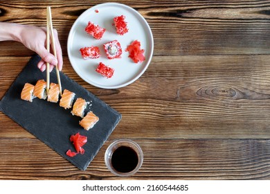 Woman Hands Holding Fish Sushi Rolls With Salmon, Red Caviar With Chopsticks On Gray Black Ceramic Serving Plate On Wooden Rustic Background Close Up. Seafood, Food Service From Restaurant Concept.